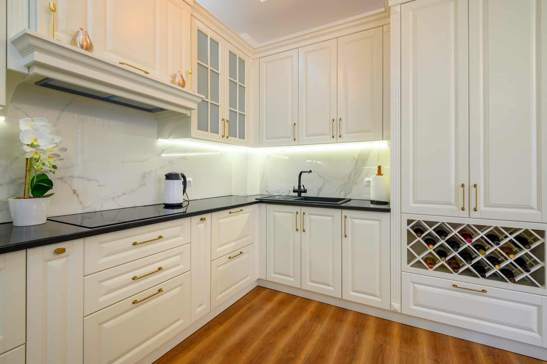 Cream Kitchen Cabinet Refinishing Cream-painted kitchen cabinetry with black countertops, brass hardware, marble backsplash, and warm wood flooring