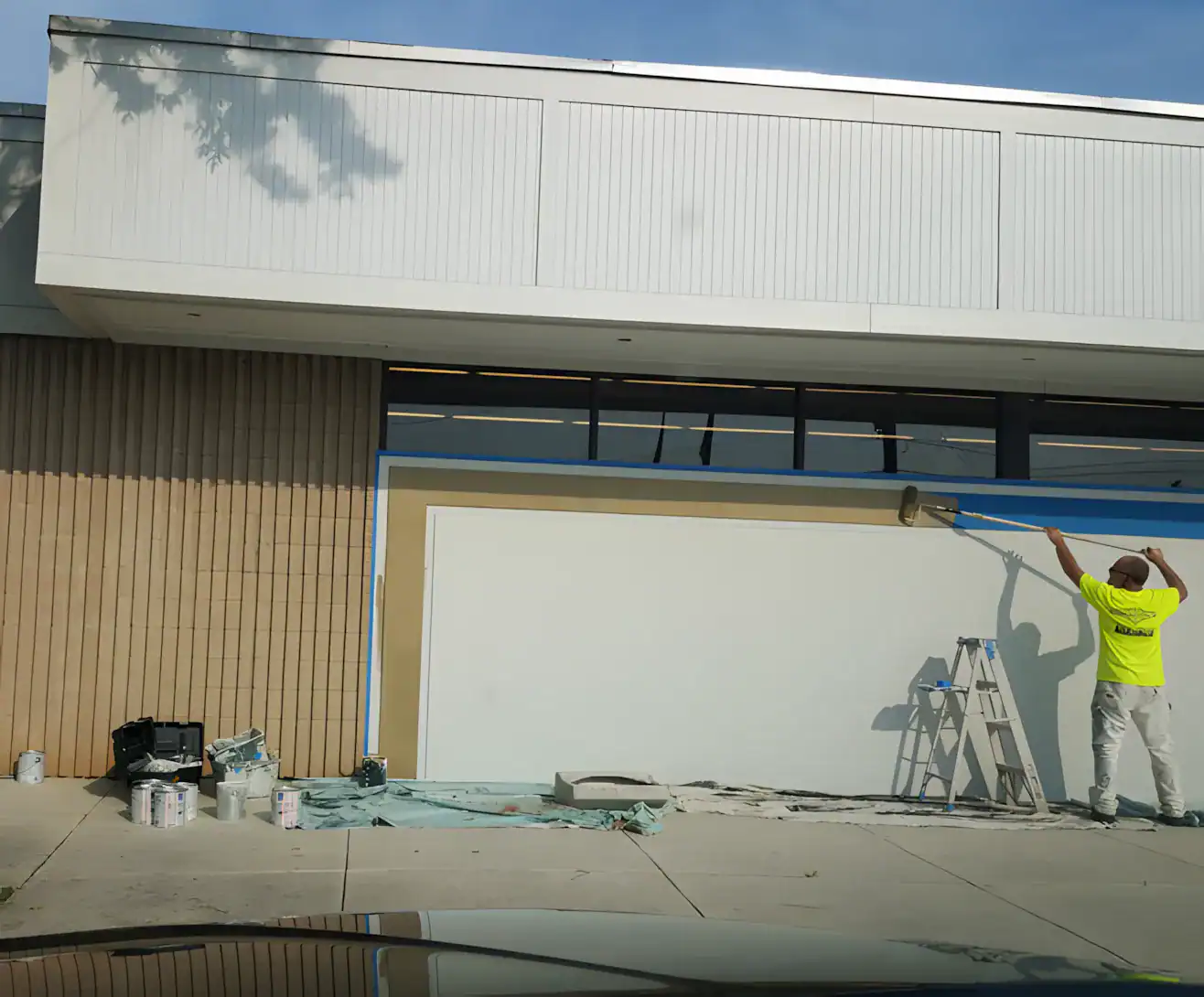Commercial Building Exterior Painting Worker in yellow safety vest painting large white commercial building facade with corrugated metal siding and blue trim