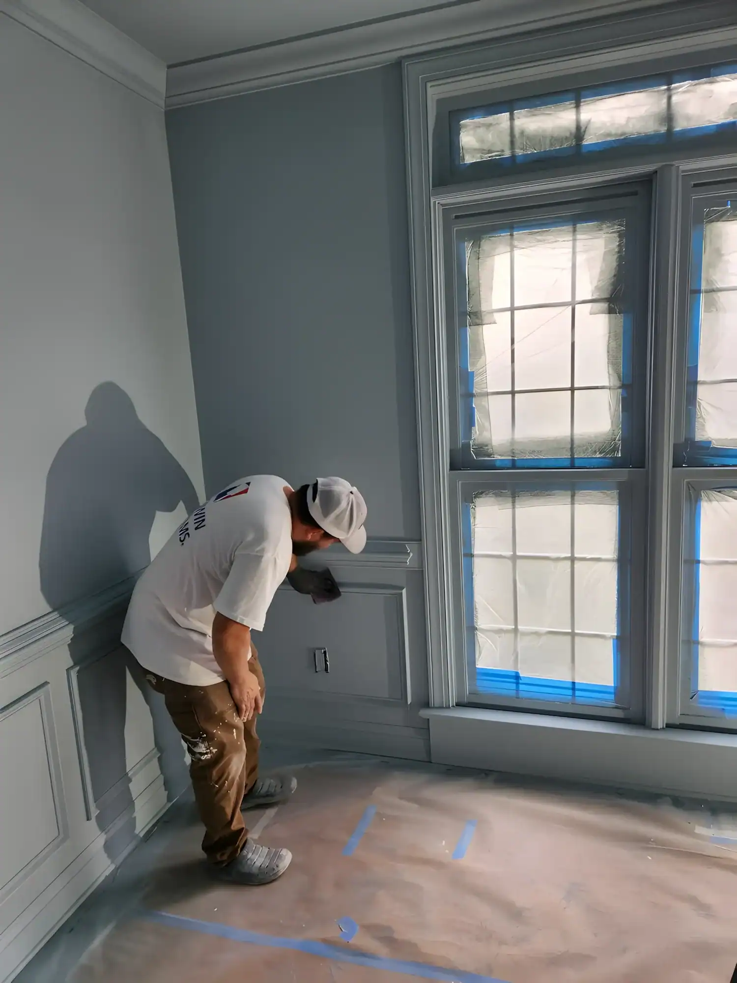 Interior Residential Painting Project Worker in white cap painting two-tone gray residential interior wall with white wainscoting and taped windows