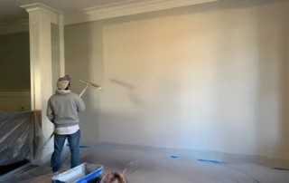 Worker painting cream-colored drywall in residential interior with blue floor protection tape and paint tray how often should you repaint interior walls